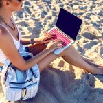 2024_07_31_consells_preventius_2024_v2 Close up image of young woman sitting on the beach and working on her laptop, backpack and hipster clothes, freelance style. Soft toned colors. Work on vacation.